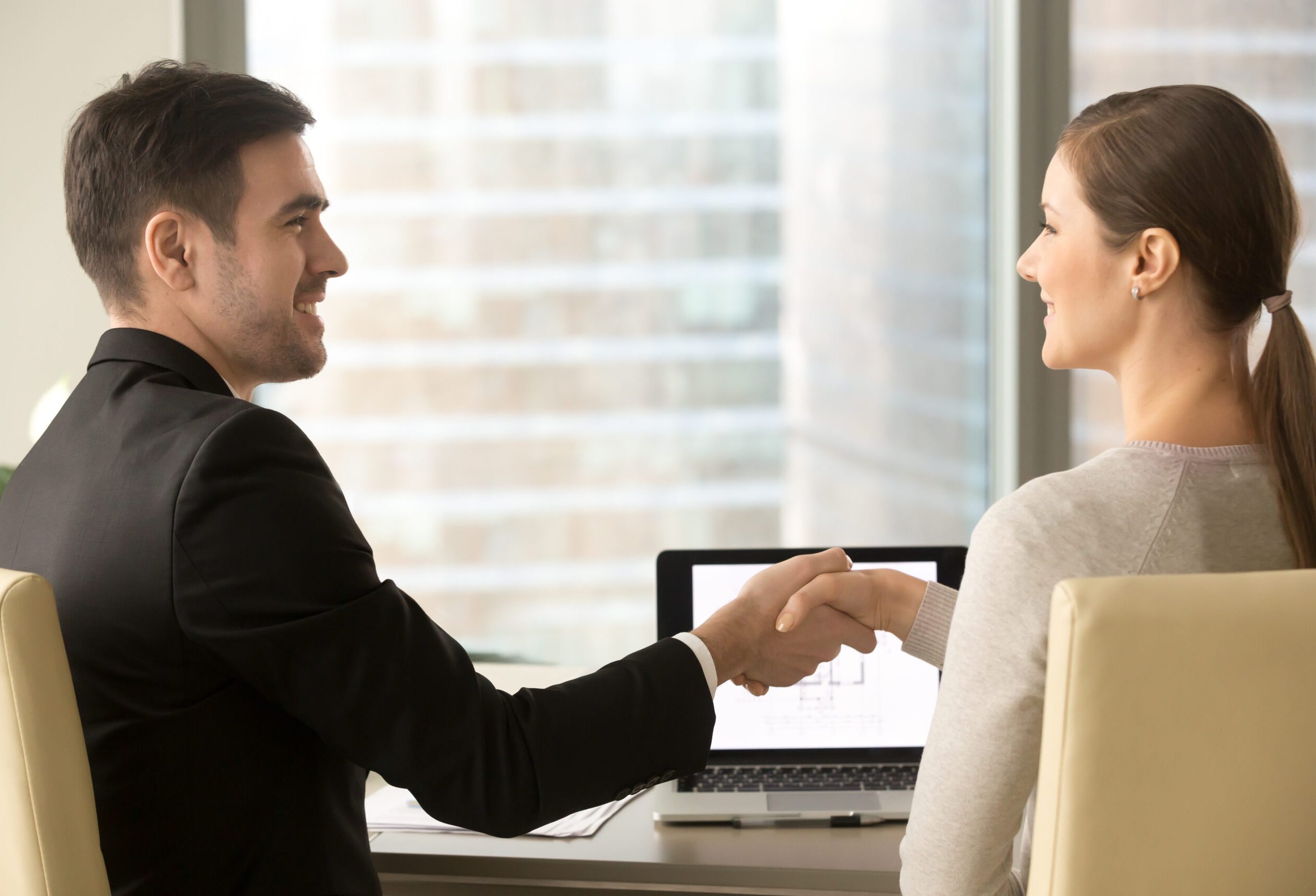 Satisfied with results of business meeting businessman and businesswoman shaking hands while sitting at desk in front of laptop. Male real estate agent congratulating female client with buying home